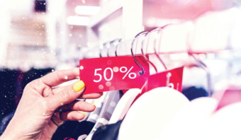 Una mano con esmalte de uñas amarillo sostiene una etiqueta de oferta roja que muestra "50%" en un perchero de ropa, resaltando las grandes promociones en la tienda.