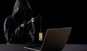Un hombre con una sudadera con capucha negra sostiene una cadena de oro frente a una computadora portátil, insinuando un posible fraude digital.