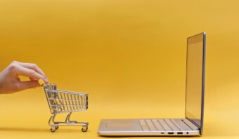 Una mano sosteniendo un carrito de compras junto a una computadora portátil sobre un fondo amarillo.