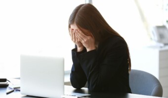 Una mujer sentada en un escritorio se cubre el rostro con las manos, mostrando signos de estrés por acoso laboral, con una computadora portátil abierta frente a ella.