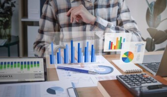 Una persona con una camisa a cuadros apuntando a la pantalla de una computadora portátil que muestra gráficos y cuadros comerciales, rodeada de documentos financieros y una calculadora en el escritorio.