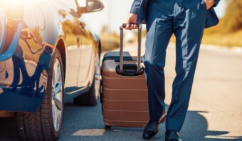 Un hombre con traje tira una maleta con ruedas junto a un auto estacionado en un día soleado, manejando la gestión de viajes, concentrándose en sus piernas y la maleta.