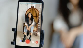 Pantalla de teléfono inteligente que muestra una grabación en vivo de un joven influencer con cabello rizado, sentado en una silla de mimbre, visto a través de la interfaz de una aplicación de cámara.