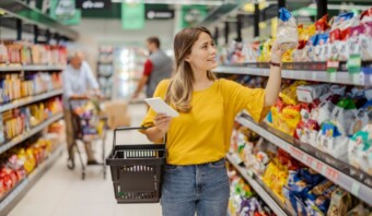 Una mujer joven con un top amarillo selecciona un producto de un estante de un supermercado mientras sostiene una canasta de compras y analiza Tendencias del consumidor mexicano.