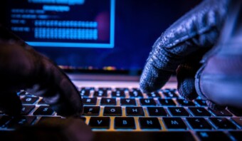 Primer plano de manos con guantes escribiendo en el teclado de una computadora portátil, iluminadas por una luz azul con el código de tráfico de bots visible en la pantalla.