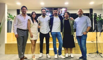 Seis personas están una al lado de la otra, sonriendo a la cámara en un espacio interior bien iluminado con una decoración moderna y plantas en macetas. La atmósfera serena rezuma elogia, lo que lo convierte en un momento perfecto para capturar.