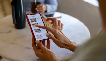 Una persona sostiene un teléfono inteligente que muestra menús digitales y usa el dedo para desplazarse. Otra persona se sienta al otro lado de la mesa.