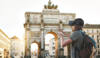 Mujer usando auriculares vr frente al arco triunfal de Siegestor en Munich, explorando contenido virtual con sus manos.