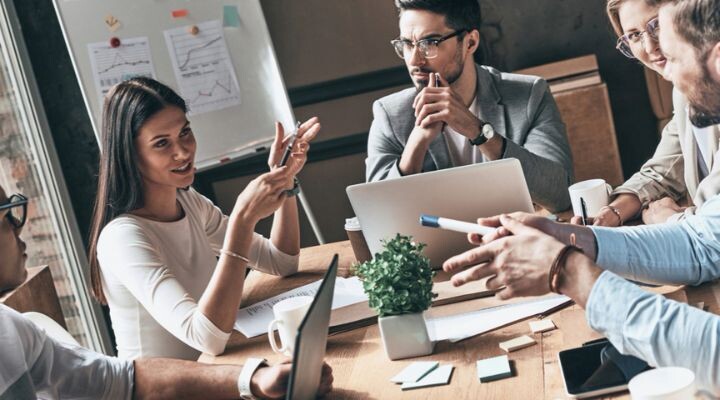 Cinco personas se sientan alrededor de una mesa con computadoras portátiles y papeles, discutiendo estrategias para manejar una crisis de marca; en un tablero al fondo se ven gráficos y notas que detallan posibles soluciones.