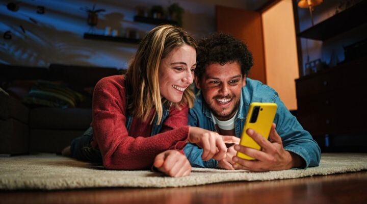 Dos personas tumbadas en un suelo moqueta en un interior, sonriendo y mirando juntas un teléfono inteligente amarillo, compartiendo un momento de ligereza incluso durante una crisis de marca.