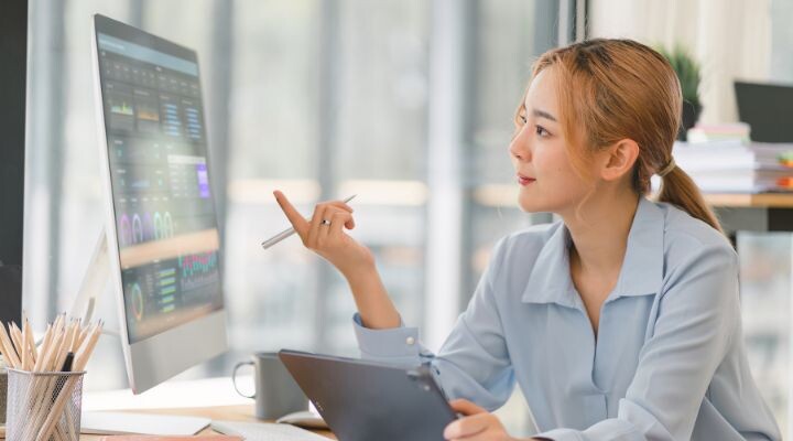 Una mujer se sienta en un escritorio frente a un gran monitor de computadora que muestra gráficos de datos, sosteniendo un lápiz y una tableta mientras analiza información impulsada por IA generativa.