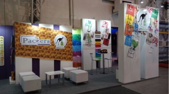 Stand de feria comercial con el logotipo de "Packters", materiales promocionales globales de marketing en exhibición, muebles de asientos blancos e imágenes de productos coloridas exhibidas en elegantes paneles blancos.