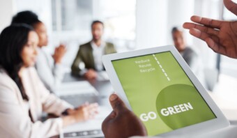 Una persona sostiene una tableta que muestra el texto "Go Green" y "Reduce, Reuse, Recicla", con cuatro personas sentadas en una mesa en un fondo borroso.