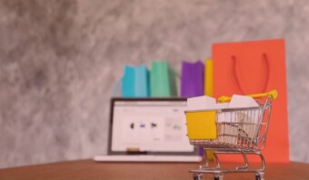 Un mini carrito de compras con cajas sobre un escritorio, una computadora portátil con un sitio de ventas en línea abierto y coloridas bolsas de compras al fondo.