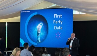 Un orador, sosteniendo un micrófono, se presenta frente a una pantalla que muestra un gráfico de "Datos de origen" en un evento de Walmart Connect. Los miembros de la audiencia están sentados y atentos en primer plano mientras la discusión profundiza en las capacidades de Walmart DSP.