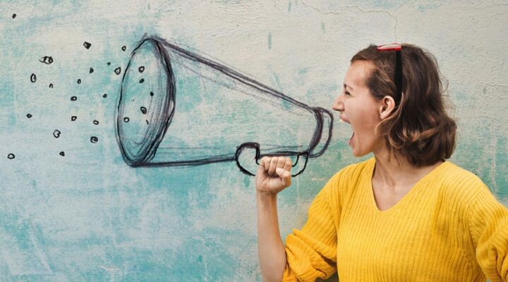 Una mujer con un suéter amarillo finge gritar en un megáfono dibujado en una pared, mientras su mano imita que lo sostiene, capturando el espíritu de una estrategia de medios creativos y visuales.