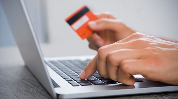 Una persona sostiene una tarjeta de crédito en una mano y escribe en el teclado de una computadora portátil con la otra, sugiriendo un pago o una compra en línea respaldada por IA en pagos.