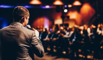 Un hombre de traje que sostiene un micrófono habla ante una audiencia sentada en un lugar espacioso y con poca luz.