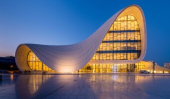 Un edificio moderno y curvilíneo con grandes ventanales se ilumina al anochecer, mostrando su sorprendente arquitectura contra un cielo azul claro.