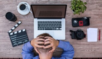 Persona sentada en un escritorio de madera con una computadora portátil, café, una cámara, una claqueta y una planta, sosteniendo su cabeza con frustración por el agotamiento.