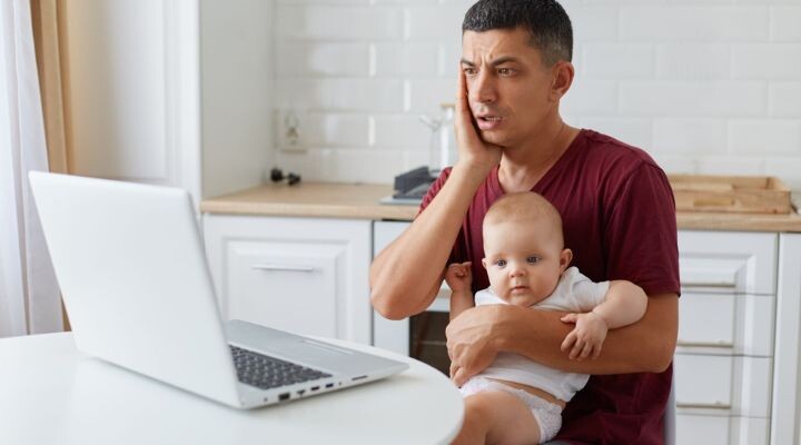 Un trabajador independiente sostiene un bebé y se sienta en una mesa de cocina, mirando la pantalla de una computadora portátil con expresión preocupada.