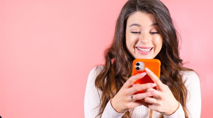 Una mujer de largo cabello castaño sonríe mientras mira su teléfono, que tiene una carcasa naranja, que encarna los alegres hábitos digitales de la generación Z, sobre un fondo rosa liso.