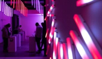 La gente interactúa con las exhibiciones en un espacio con poca luz, bañado por luces violetas y rosas, con tubos de luz verticales colgando del techo, un ejemplo perfecto de la industria creativa en acción.