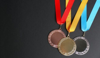 Tres medallas con cintas de colores (rojo, amarillo y azul) se muestran sobre un fondo negro. Las medallas parecen ser de bronce, oro y plata, que recuerdan a las que se otorgan en los juegos olímpicos.