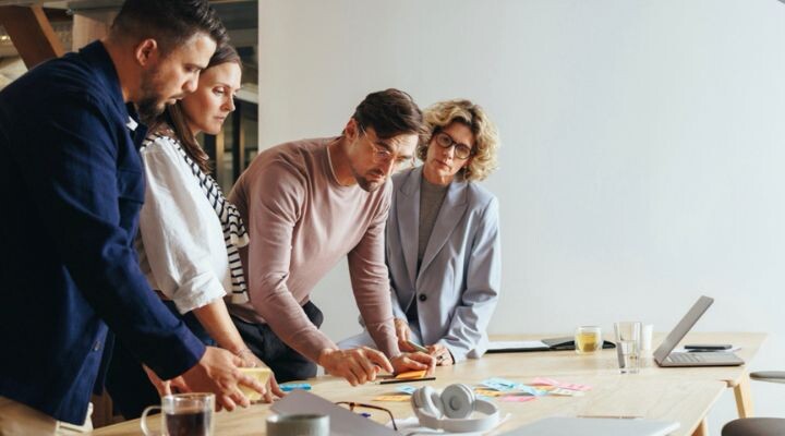 Cuatro personas están alrededor de una mesa en una oficina, colaborando en documentos y notas adhesivas, con una computadora portátil y auriculares cerca, fomentando un ambiente de innovación.