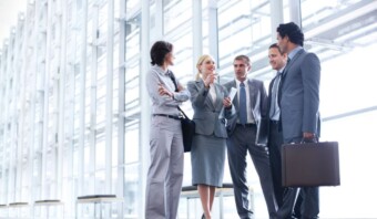 Cinco profesionales de negocios se encuentran en el pasillo de una oficina moderna, conversando. Tres hombres y dos mujeres visten traje y un hombre sostiene un maletín. Parecen estar discutiendo estrategias de relaciones públicas.