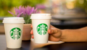Dos tazas de café Starbucks sobre una mesa, una sostenida por una mano. Al fondo se ve una planta en maceta.