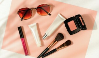 Un par de gafas de sol, un brillo de labios, dos brochas de maquillaje, dos tubos y un kit de rubor compacto están dispuestos sobre una superficie rosa, creando una escena de belleza natural.