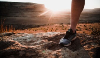 Una persona que lleva zapatillas para correr se encuentra en un terreno rocoso con un paisaje desértico y una puesta de sol de fondo.