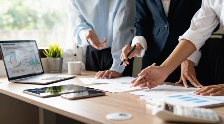 Un equipo de cuatro profesionales vestidos de negocios discuten cuadros y gráficos financieros en un escritorio con una computadora portátil, una tableta y papeleo durante una reunión.