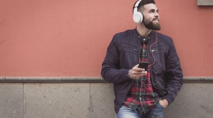 Un hombre con auriculares y una chaqueta se apoya contra una pared roja, sosteniendo un teléfono inteligente y mirando hacia un lado, como si sintonizara la radio para escuchar su música favorita.