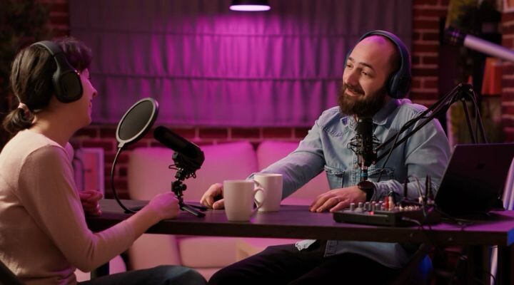 Dos personas con auriculares se sientan en una mesa con micrófonos y tazas de café, grabando un podcast en un estudio con iluminación violeta que emite vibraciones clásicas de la radio.