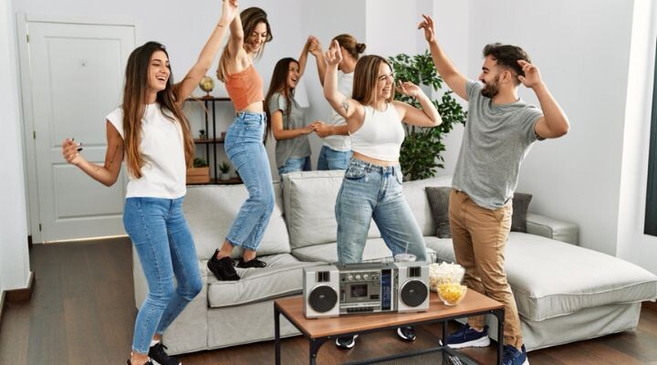 Cinco jóvenes bailan y sonríen en una sala de estar junto a una mesa con radio, palomitas de maíz y bocadillos.