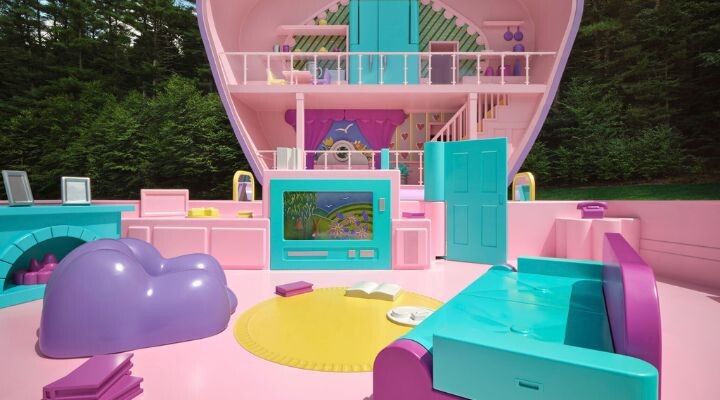 Un interior de casa de muñecas de tamaño natural, en colores pastel, inspirado en Polly Pocket, con muebles de color rosa, verde azulado y morado, una alfombra amarilla y un libro abierto, ambientado al aire libre con árboles de fondo.