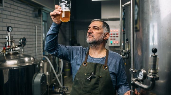 Un hombre con delantal examina un vaso de cerveza dentro de una cervecería, rodeado de tanques de acero inoxidable y equipos de elaboración, reflejando las últimas tendencias en la elaboración artesanal.