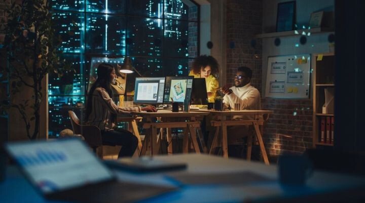 Tres personas trabajan hasta altas horas de la noche en una oficina moderna, discutiendo ideas publicitarias mientras usan computadoras, con las luces de la ciudad visibles a través de grandes ventanas.