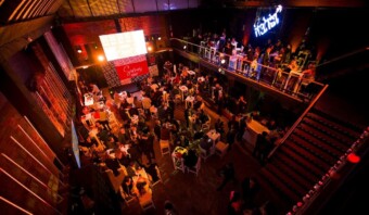 Un gran espacio interior para eventos con gente socializando en mesas, una pantalla brillante que muestra texto y un letrero de neón sobre un balcón en el segundo piso. El ambiente es animado con una iluminación cálida.