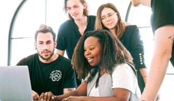 Un grupo de cinco personas, tres hombres y dos mujeres, se reúnen alrededor de una computadora portátil con expresiones concentradas. La persona que está al frente escribe en el teclado mientras otros observan, mostrando una efectiva atracción de talento en acción.