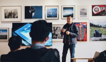 Una persona se pone de pie y utiliza la narración para involucrar a una audiencia, con una pantalla grande y fotografías enmarcadas en la pared detrás de ella.
