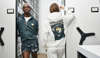 Dos personas se encuentran entre los almacenes. Una, con un atuendo con motivos de la NASA, mira a la cámara. La otra, de espaldas, lleva una sudadera con capucha de Reebok y hace un gesto con el brazo levantado.