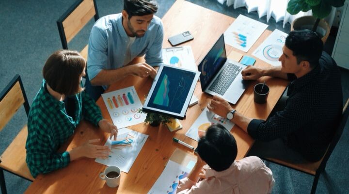 Cuatro personas en una sala de reuniones están sentadas alrededor de una mesa de madera y colaboran con documentos, gráficos y computadoras portátiles. Una persona señala un gráfico en una tableta, optimizando el presupuesto mientras participa en una planificación detallada de los medios.