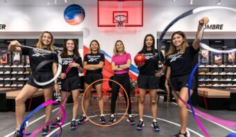Un grupo de seis mujeres, cinco de ellas con equipamiento deportivo en la mano, se encuentran en una tienda Reebok en Mundo E, con una exposición de zapatos al fondo.
