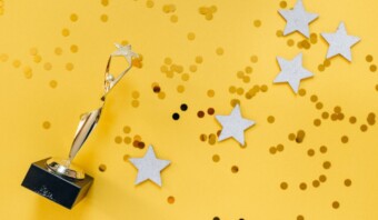 Un trofeo dorado con una estrella en la parte superior se encuentra sobre una superficie amarilla salpicada de estrellas blancas y puntos de confeti dorados, encarnando el espíritu de prevmios creativos.