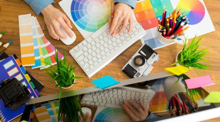 Persona que usa un mouse y un teclado de computadora en un escritorio con muestras de color, lápices de colores, cámara, plantas y un monitor, mostrando un espacio de trabajo publicitario creativo.