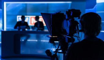 Una persona opera una cámara de televisión en un estudio, enfocando a dos presentadores de noticias sentados en un escritorio en el fondo, con un escenario iluminado en azul que recuerda a los que se ven en los reality shows populares.