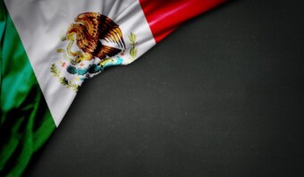 La bandera mexicana, que muestra rayas verticales verdes, blancas y rojas con un escudo de armas que incluye un águila y un cactus (los símbolos patrios del país), cubre parcialmente una superficie oscura.